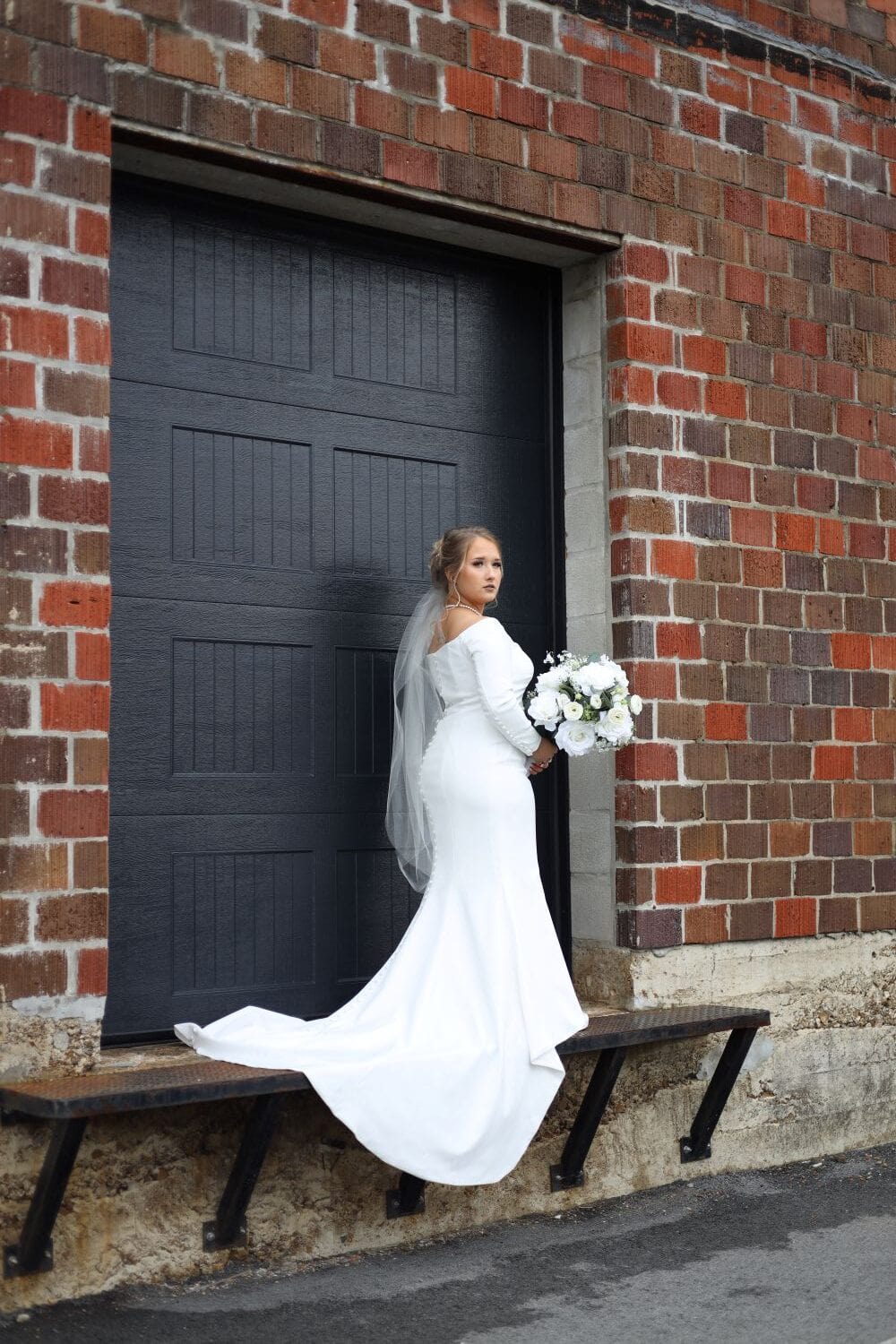 Bride in white wedding dress holding bouquet outside urban brick building.
