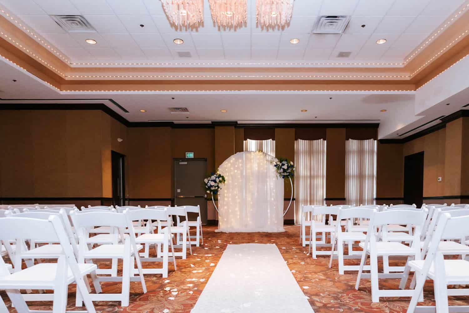 Wedding ceremony setup with white chairs and floral arch in a decorated hall.