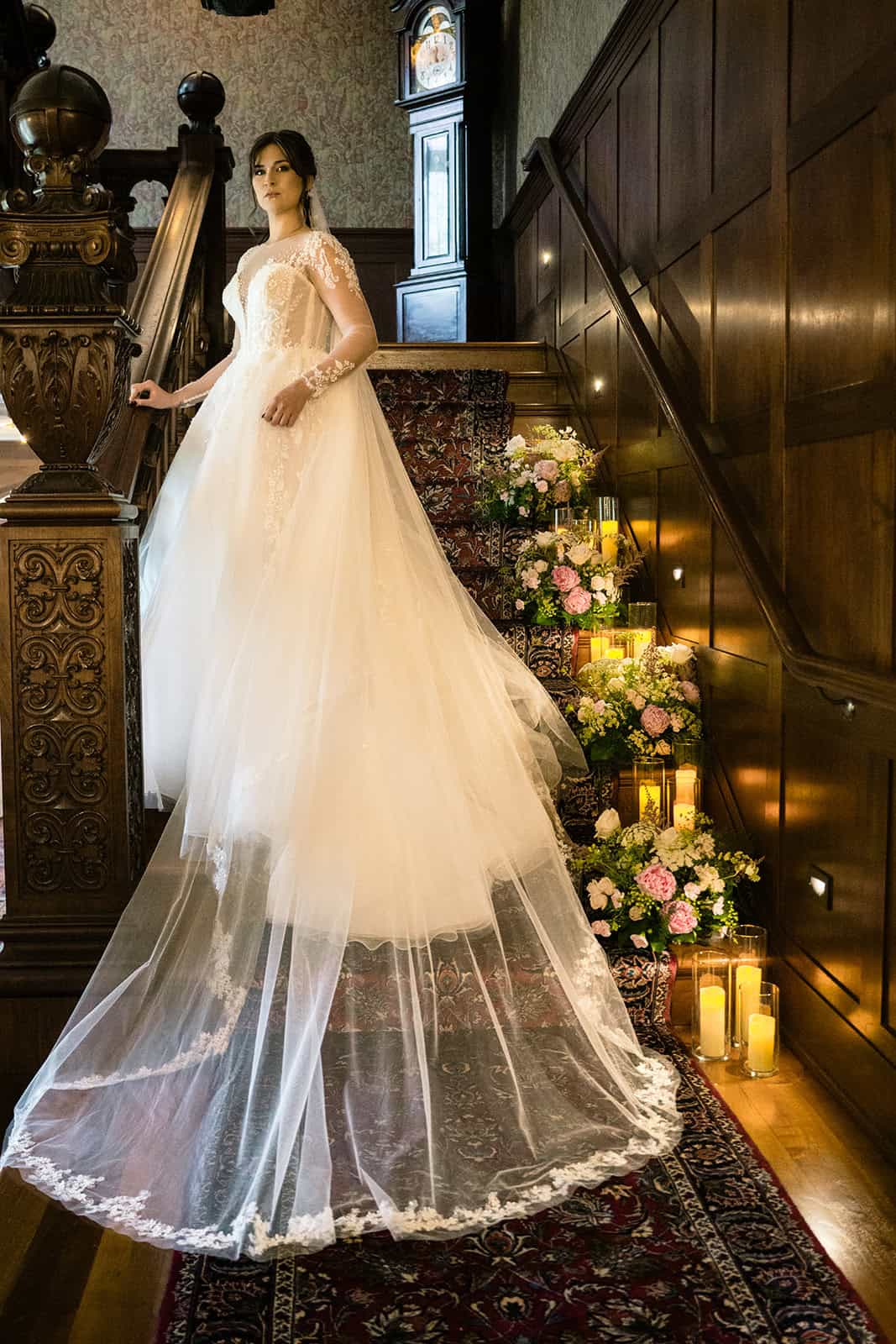 Bride in a white wedding gown on staircase with floral decor and candles.